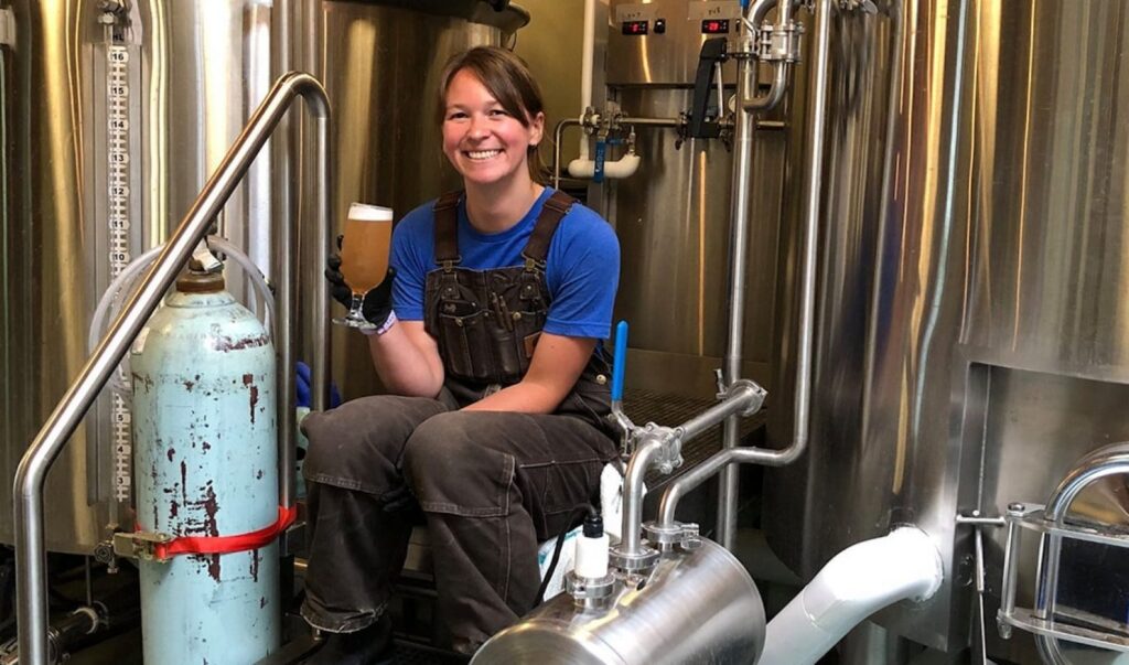 A woman sits on the stairs of a brewing system as she smiles and has a tall glass of beer in her hand.