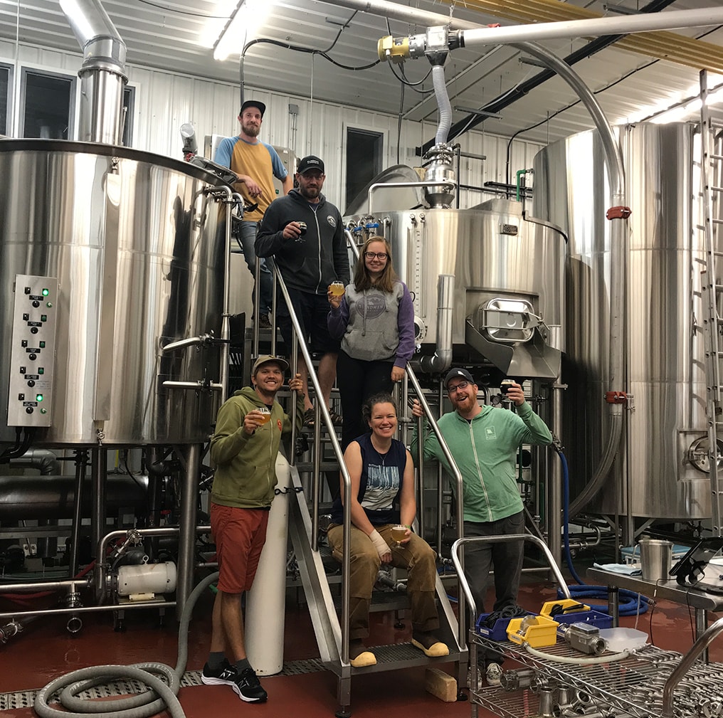 A group of people stand on stairs holding a glass of beer in front of 30-100 BBL brewing tanks.