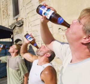 Three men have their heads tilted back all drinking a bottle of beer.