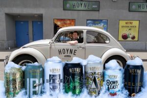 A off white volkswagen beetle with the Hoyne Brewing Company logo on the side of it. Seven cans of Hoyne beer line the front with ice all around the cans.