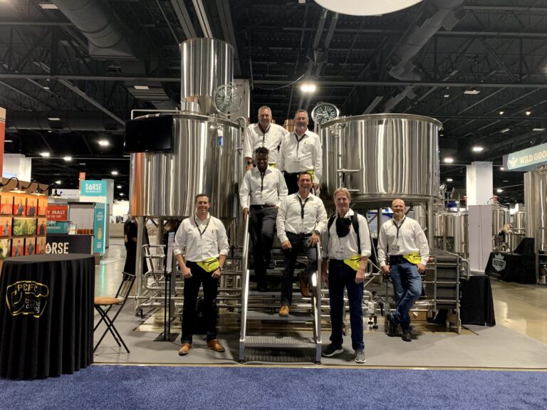 A group of men in white shirts and yellow belt bags stand on the stairs going up to two craft brewing tanks.