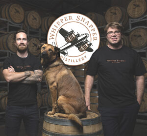 Whipper Snapper Distillery logo hangs behind two men standing smiling as their dog sits on a barrel.