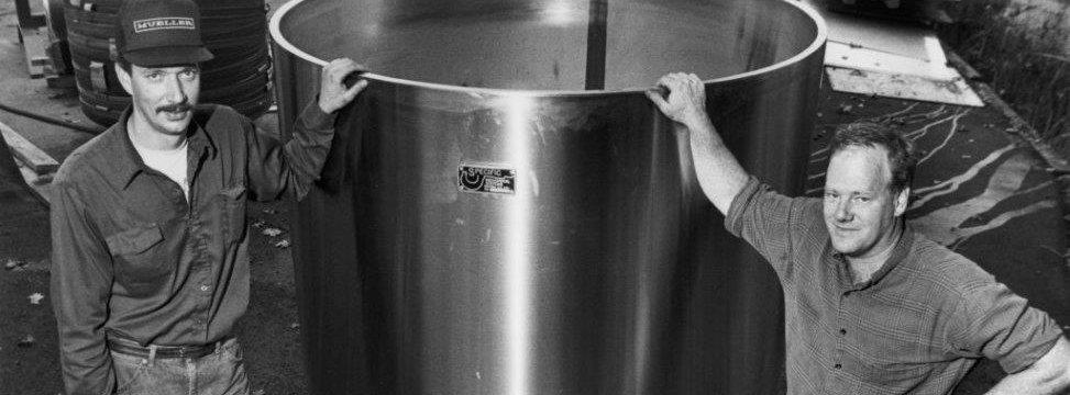 A black and white photograph of two founders each have one hand on a brewing tank smiling.