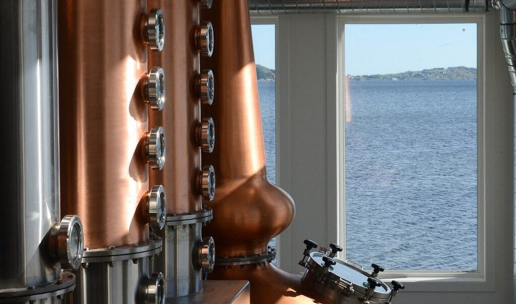 A close up of two distilling columns with the ocean in the background outside the window.